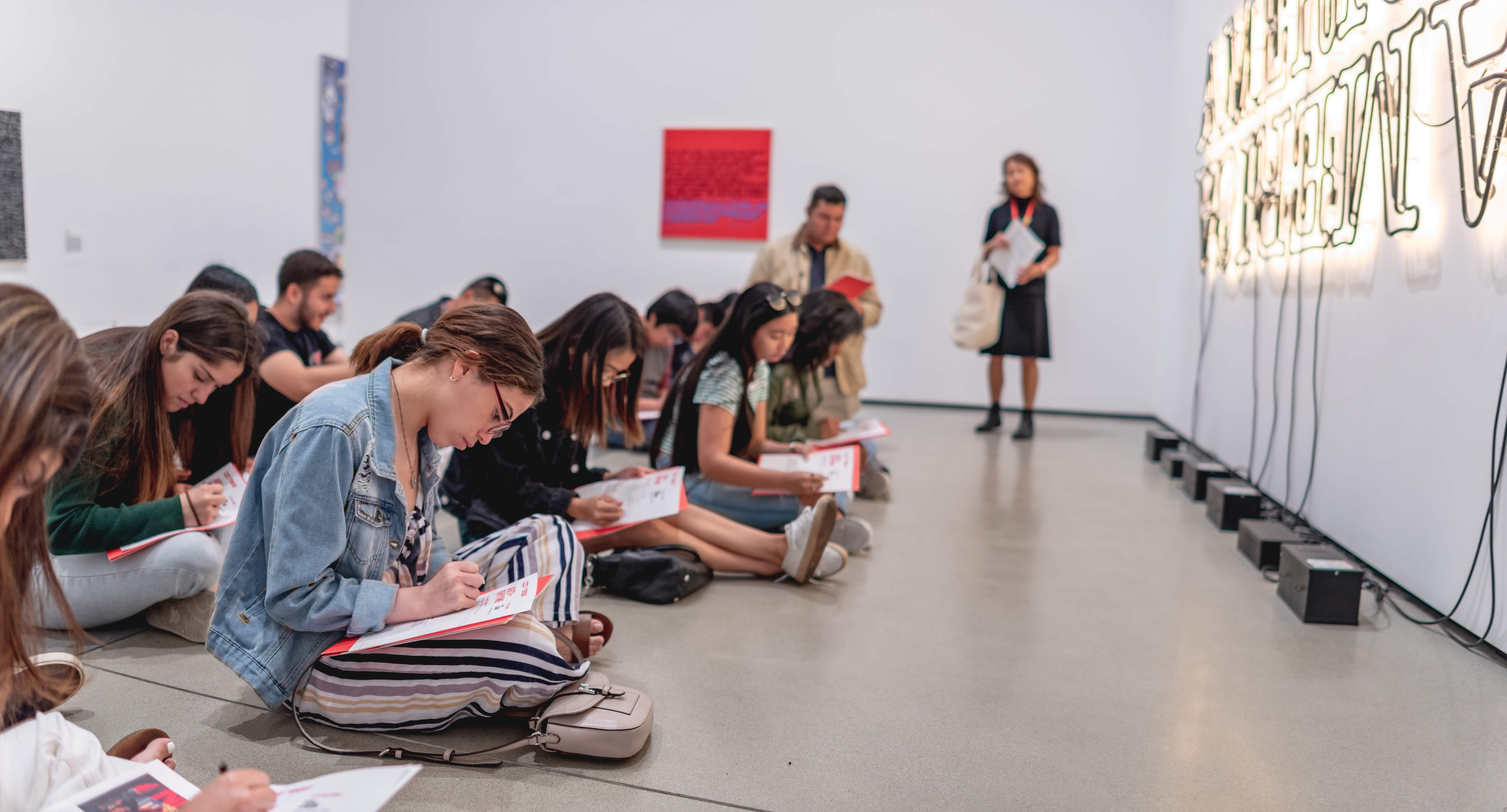 Teenagers sitting on the floor of The Broad writing on a sheet of paper looking at artwork