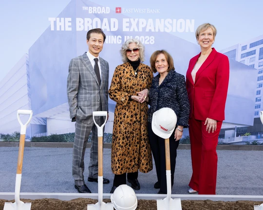 A group, one man and three women, in suits, standing behind shovels dug into a patch of dirt