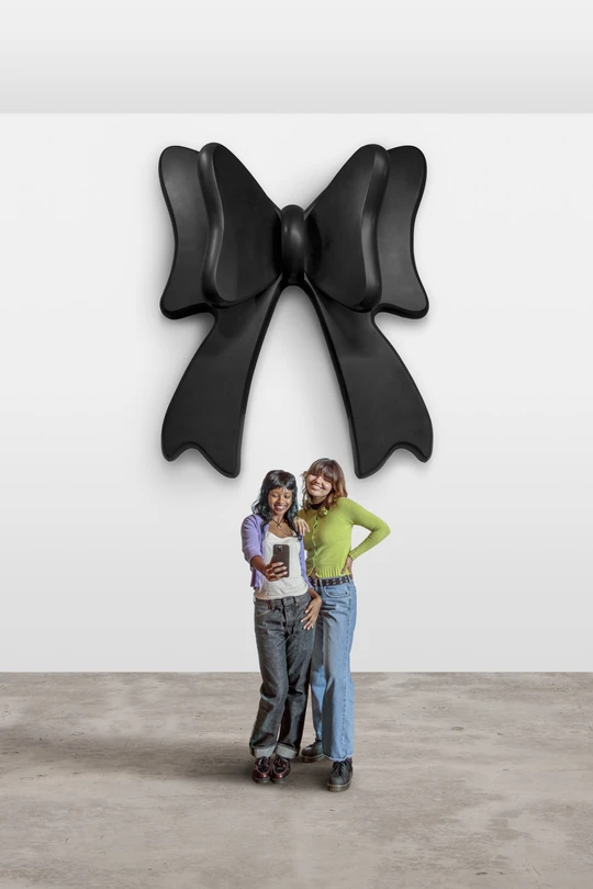 Two young women taking a selfie in front of Robert Therrien's oversized bow sculpture which is black and hanging on a wall in back of them